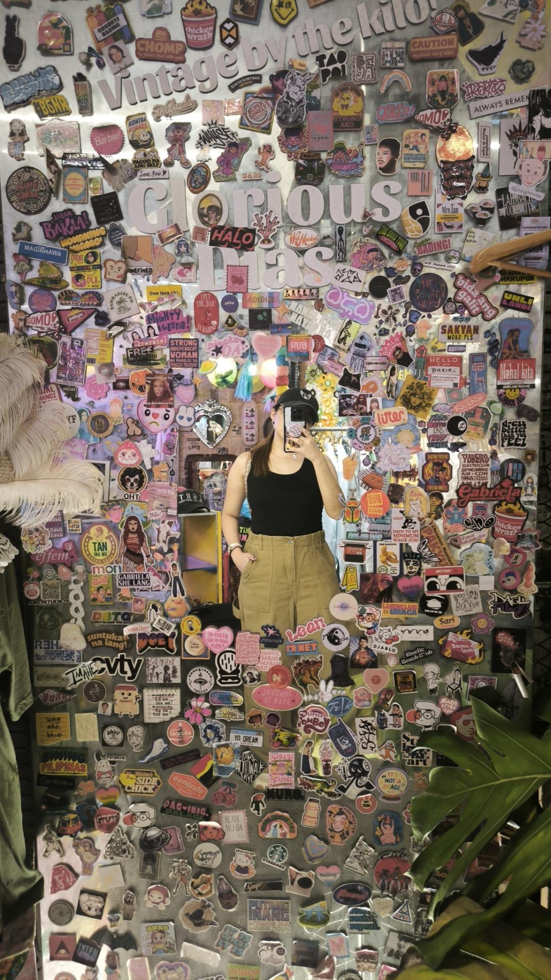 mirror selfie woman in black top and khaki jeans with hat, mirror is completely surrounded by stickers